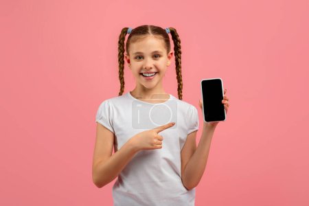 A happy girl points at her smartphone screen, looking at the camera with a smile on a pink background