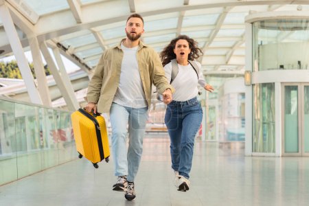 Téléchargez les photos : Un jeune couple se précipite à travers un terminal de l'aéroport, l'urgence gravée sur leurs visages, les bagages en remorque. - en image libre de droit