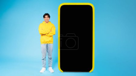 Photo for A smiling young Asian guy with crossed arms stands beside a large smartphone prop with a blank screen mockup - Royalty Free Image