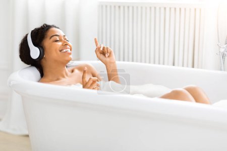 Téléchargez les photos : Femme afro-américaine bénéficie d'un bain paisible avec écouteurs, signalant un moment de détente au milieu d'une vie occupée - en image libre de droit