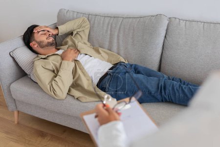 Photo for A man in casual attire is lying on a couch with his hand on his forehead, possibly in a deep thought or distress, indicative of a therapy or counseling session - Royalty Free Image