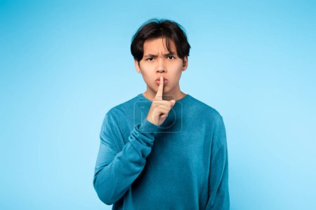 Photo for Asian guy requests silence by placing a finger on his lips with a serious expression on a blue background - Royalty Free Image