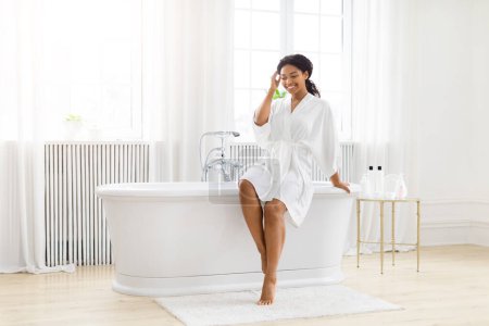 Téléchargez les photos : Une jeune afro-américaine souriante profite d'une matinée ensoleillée, assise dans une élégante baignoire dans une salle de bain ensoleillée - en image libre de droit