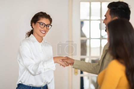 A therapist woman in glasses extends a friendly handshake to a couple in a welcoming gesture during a meeting
