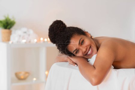 Téléchargez les photos : Femme afro-américaine avec un sourire ravi repose sur une table de massage spa, les mains doucement placées sous son menton - en image libre de droit