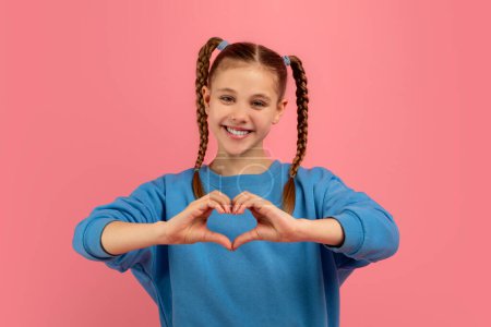 Young girl in blue top creates a heart shape with her hands, symbolizing love and affection on pink background