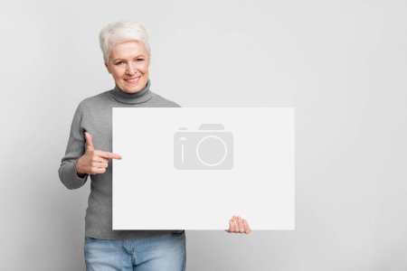 Foto de Una anciana europea sonriente y anciana sostiene alegremente una gran pizarra blanca en blanco, ideal para anuncios de s3niorlife - Imagen libre de derechos