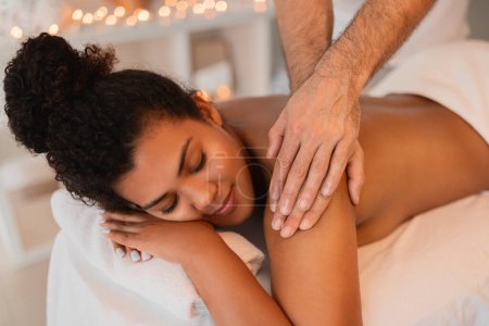Téléchargez les photos : Une femme afro-américaine détendue recevant un massage doux des épaules dans un environnement spa serein avec éclairage ambiant - en image libre de droit