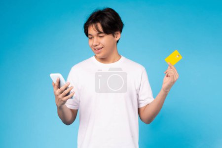 Téléchargez les photos : Jeune Asiatique gars tient un smartphone et montrant une carte de crédit, suggérant achats en ligne ou de paiement - en image libre de droit