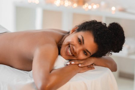 Foto de Una alegre mujer afroamericana con el pelo rizado sonríe tumbada cómodamente sobre una mesa de masaje blanca en un entorno de spa - Imagen libre de derechos