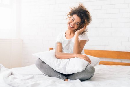 Foto de Buenos días. Mujer positiva sentada en la cama con almohada y sonriendo a la cámara - Imagen libre de derechos