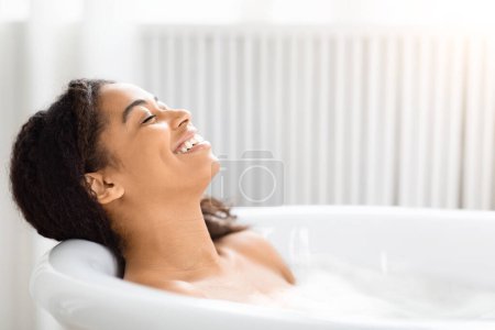 Téléchargez les photos : Une femme afro-américaine joyeuse avec un sourire radieux, profitant d'un bain moussant relaxant dans un cadre lumineux de salle de bain - en image libre de droit