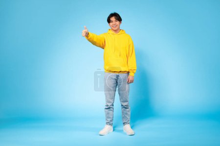 Téléchargez les photos : Un jeune Asiatique ravi dans un sweat-shirt jaune donne un pouce vers le haut, exprimant son bonheur sur un fond bleu - en image libre de droit