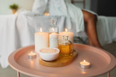 Téléchargez les photos : Cadre spa relaxant avec des bougies allumées, de l'huile de massage et un bol sur une table d'appoint rose au-dessus d'une dame noire en peignoir blanc - en image libre de droit