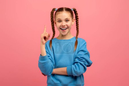 Téléchargez les photos : Une jeune fille joyeuse aux cheveux tressés pointe vers le haut, debout sur un fond rose avec un grand sourire - en image libre de droit