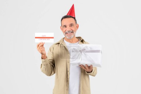 Téléchargez les photos : Un homme âgé pose avec un chapeau de fête, un certificat-cadeau et une boîte-cadeau blanche, incarnant la célébration et la générosité - en image libre de droit