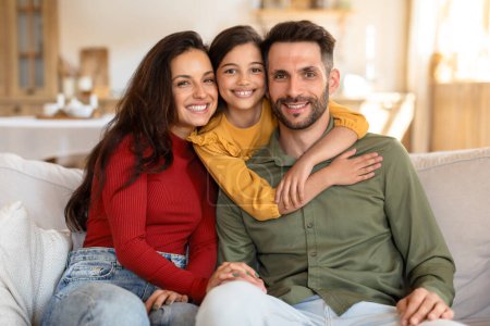 Foto de Retrato familiar cálido con los padres y la hija abrazando, compartiendo un momento de afecto en el sofá de su casa - Imagen libre de derechos
