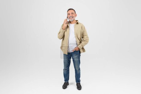 Un anciano con barba sonríe mientras habla por teléfono, vestido casualmente con chaqueta caqui y jeans, de pie sobre un fondo blanco