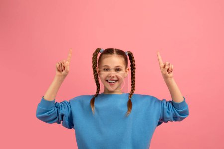 Cheerful young girl wearing a blue sweater with braided hair joyfully pointing upwards on a pink background