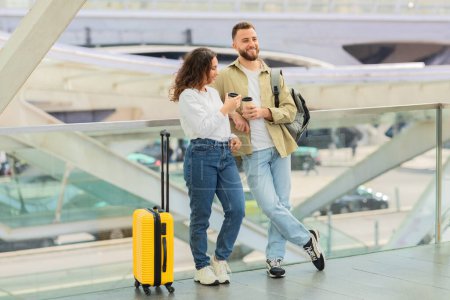 Foto de Hombre y mujer con café para llevar esperando vuelo en el aeropuerto, sonriente pareja joven con equipaje de pie en la terminal, charlando y riendo, emocionado por el próximo viaje - Imagen libre de derechos