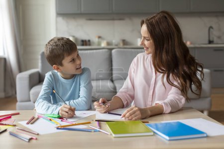 Téléchargez les photos : Une mère en chemise rose est tournée vers son fils, conversant et dessinant avec des crayons de couleur sur papier, indiquant un moment éducatif ou collant à la maison - en image libre de droit