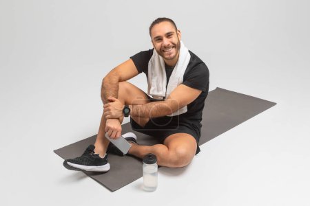 Hombre sonriente sentado casualmente en una alfombra de gimnasio con un teléfono, botella de agua y toalla, estudio gris, tienen descanso