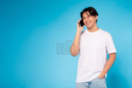 A cheerful young Asian guy standing and having a pleasant conversation on his smartphone, looking at copy space