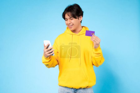 Téléchargez les photos : Jeune Asiatique gars dans un sweat à capuche jaune regardant le téléphone avec une carte de crédit, symbolisant pratique achats en ligne - en image libre de droit