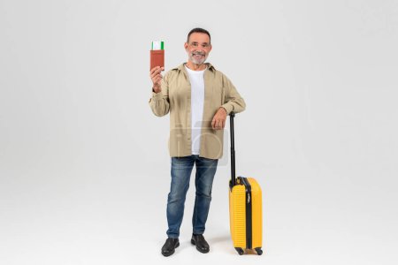 A smiling older man in casual clothing stands with a yellow suitcase and a passport, indicating he is ready for travel or vacation