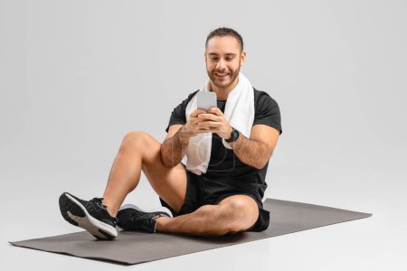 Téléchargez les photos : Homme joyeux utilisant un smartphone assis sur un tapis de fitness dans un cadre de studio d'exercice, belle application - en image libre de droit