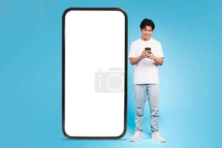 Foto de Un joven asiático se para junto a una pantalla de teléfono de gran tamaño con una pantalla en blanco, ideal para contenido publicitario - Imagen libre de derechos