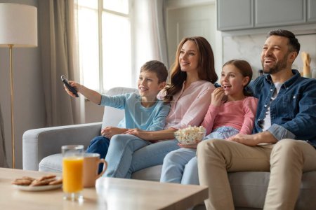 Famille joyeuse avec deux enfants assis sur un canapé et appuyant sur la télécommande tout en regardant la télévision à la maison