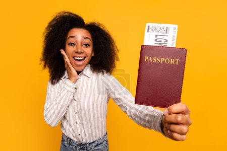Téléchargez les photos : Jeune femme afro-américaine avec une expression surprise tenant son passeport avec un billet d'avion sur un fond jaune - en image libre de droit
