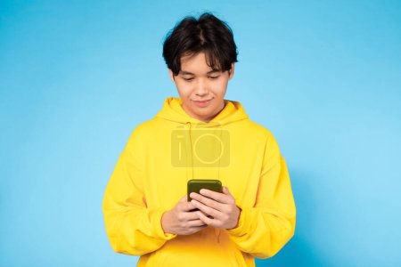 A focused young Asian guy in yellow sweatwear deeply engaged with texting on his phone on a blue background