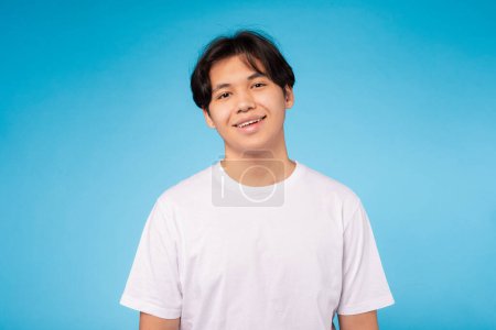 Téléchargez les photos : Un jeune Asiatique heureux avec un sourire chaleureux, portant un t-shirt blanc décontracté sur un fond bleu, gros plan - en image libre de droit