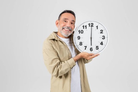 A cheerful senior man with a beard smiling and holding a large analog wall clock in front of a white background