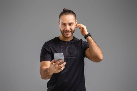 Photo for A smiling man holds a smartphone and press on earbud, accept phone call against a gray studio background - Royalty Free Image