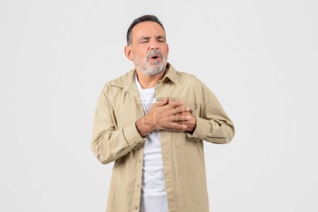 A senior man stands against a white background, clutching his chest in pain with a pained expression, possibly indicating a medical emergency like a heart attack