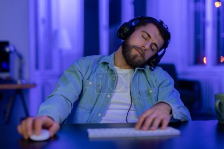 Téléchargez les photos : Épuisé gars gamer s'endormir tandis que le jeu en ligne sur son ordinateur personnel dans une pièce éclairée par des néons - en image libre de droit