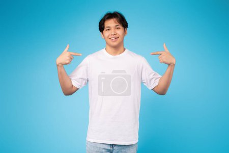 Selbstbewusster asiatischer Teenager in weißem Hemd, der vor einem soliden blauen Studiohintergrund auf sich selbst zeigt