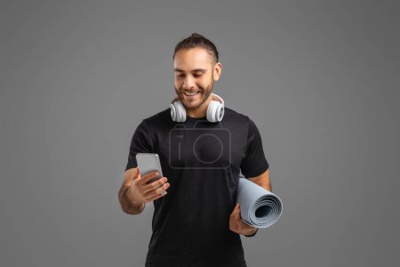 Hombre sonriente comprueba su teléfono inteligente mientras sostiene una estera de yoga, posiblemente revisando una aplicación de fitness sobre fondo gris