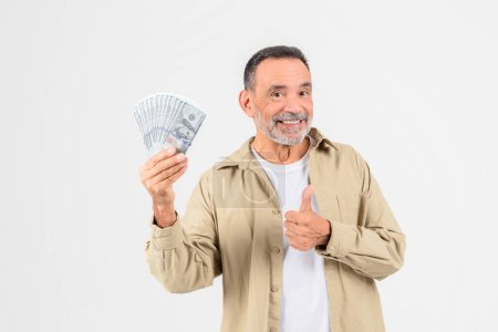 Téléchargez les photos : Un homme âgé souriant en tenue décontractée tient un fan d'argent dans une main et donne un pouce vers le haut, signalant le succès financier ou des économies - en image libre de droit