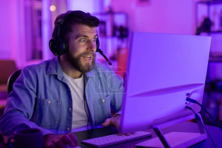Téléchargez les photos : Homme avec écouteurs intensément concentré sur un écran d'ordinateur, entouré d'une pièce maussade éclairée bleue à la maison - en image libre de droit