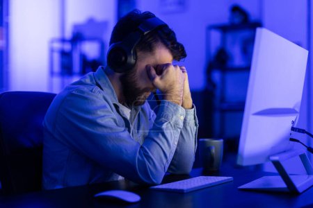 Un hombre estresado con auriculares sostiene su cabeza desesperada en un escritorio de computadora, rodeado de luz azul oscura