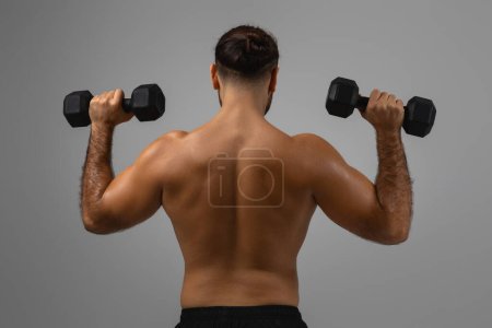 Photo for A muscular mans back is on display as he performs an exercise with black dumbbells against a grey backdrop - Royalty Free Image