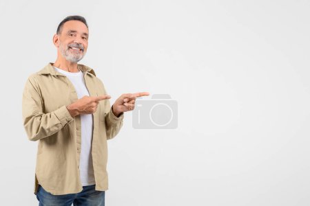 Téléchargez les photos : Un homme âgé attrayant avec un petit sourire pointe de côté avec les deux mains, indiquant quelque chose d'invisible sur fond blanc - en image libre de droit