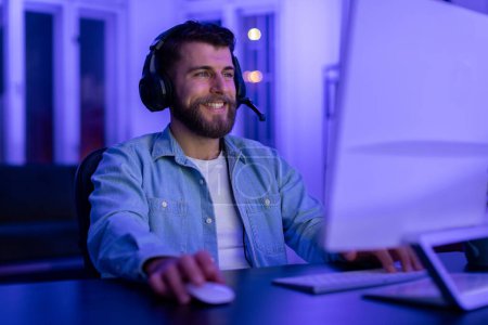 Foto de Contenido hombre barbudo gamer con auriculares disfruta de los juegos en una habitación iluminada por neón con enfoque dedicado en casa - Imagen libre de derechos