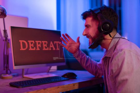 Téléchargez les photos : Homme barbu frustré gamer réagit à un message de défaite sur son écran d'ordinateur dans une pièce sombre et éclairée - en image libre de droit
