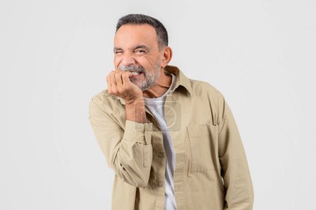 Un alegre caballero de edad en una camisa beige casual sonriendo con una mano apoyada en su barbilla, sobre un fondo gris