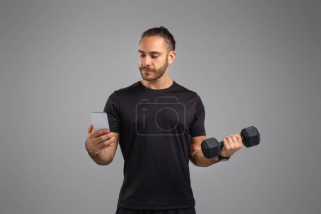A man lifts a dumbbell while simultaneously checking his smartphone, illustrating fitness mobile app